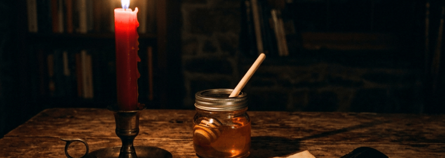 Red candle burning in brass holder, jar of honey with dipper, and handwritten note with quill on wooden table