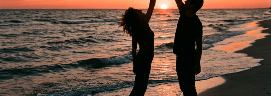 Silhouettes of a man and woman reaching towards the setting sun on a beach