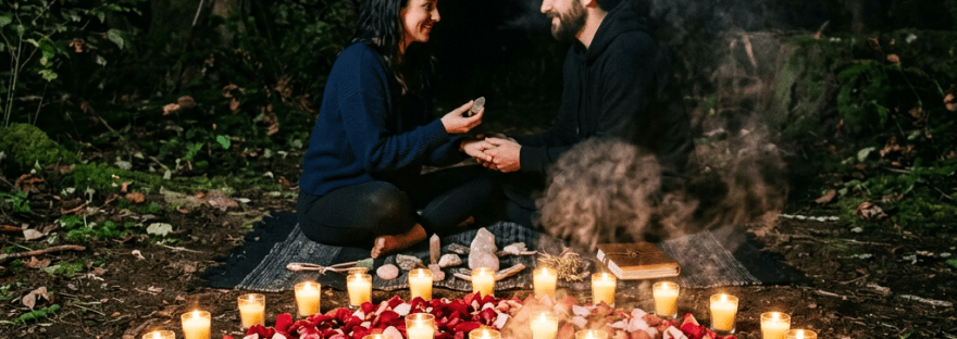 Couple sitting on a blanket outdoors at night surrounded by lit candles and rose petals in a circular pattern