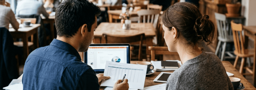 Two people sitting at a wooden table, reviewing printed documents and a laptop