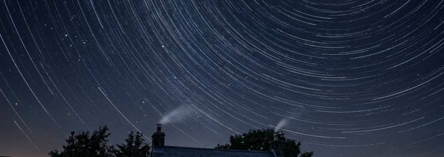Stone cottage glowing beneath circular star trails and a full moon