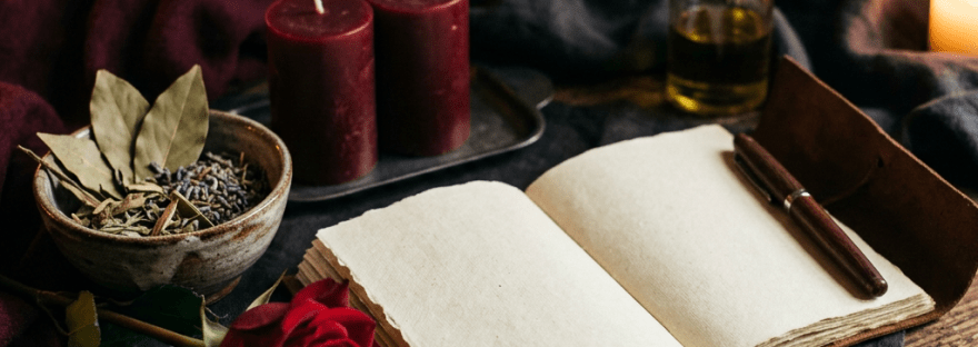 Open blank journal beside candles, herbs, oil bottle, pen, and red rose